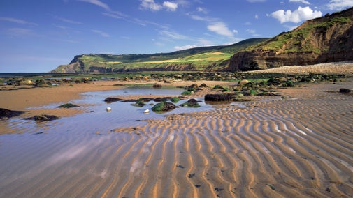 A sunlit beach and cliffs at Boggle Hole near Robin Hood's Bay with a view towards Ravenscar. A wide expanse of rippled sand dotted with pools and rocks in the foreground, with cliffs on the right, curving into a rocky headland topped with green fields in the distance.